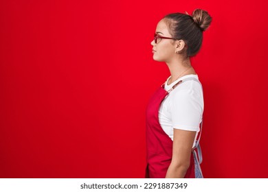 Young hispanic woman wearing waitress apron over red background looking to side, relax profile pose with natural face with confident smile. 
