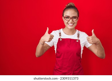 Young hispanic woman wearing waitress apron over red background success sign doing positive gesture with hand, thumbs up smiling and happy. cheerful expression and winner gesture.  - Powered by Shutterstock