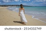 Young hispanic woman in a white dress walks along the sandy beach of pescoluse in puglia, italy, enjoying the scenic salento coastline under a bright blue sky.