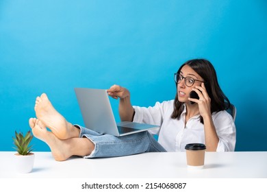 Young Hispanic Woman Working Feet On Desk Isolated On Blue Background. Boss Giving Orders On Phone.