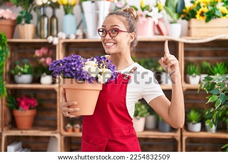 Young hispanic woman working at florist shop holding plant smiling with an idea or question pointing finger with happy face, number one 