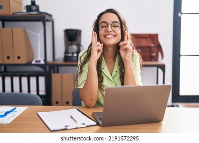 Young hispanic woman working at the office wearing glasses gesturing finger crossed smiling with hope and eyes closed. luck and superstitious concept.  - Powered by Shutterstock