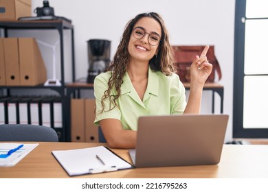 Young Hispanic Woman Working At The Office Wearing Glasses With A Big Smile On Face, Pointing With Hand And Finger To The Side Looking At The Camera. 