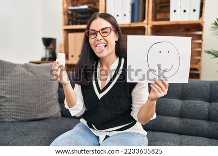 Young hispanic woman working on depression holding happy emotion on paper and pills sticking tongue out happy with funny expression. 