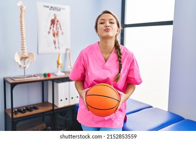 Young hispanic woman working at physiotherapy clinic holding basketball ball looking at the camera blowing a kiss being lovely and sexy. love expression. 