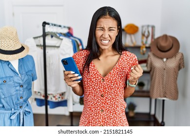 Young hispanic woman working at retail boutique using smartphone angry and mad screaming frustrated and furious, shouting with anger. rage and aggressive concept. 