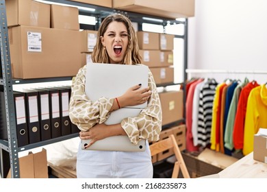 Young hispanic woman working at retail boutique holding laptop angry and mad screaming frustrated and furious, shouting with anger. rage and aggressive concept. 