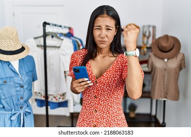 Young hispanic woman working at retail boutique using smartphone angry and mad raising fist frustrated and furious while shouting with anger. rage and aggressive concept. 