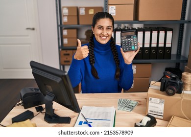 Young Hispanic Woman Working At Small Business Ecommerce Holding Calculator Smiling Happy And Positive, Thumb Up Doing Excellent And Approval Sign 