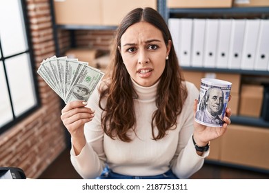 Young Hispanic Woman Working At Small Business Ecommerce Holding Money And Piggy Bank Clueless And Confused Expression. Doubt Concept. 