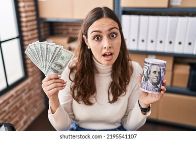 Young Hispanic Woman Working At Small Business Ecommerce Holding Money And Piggy Bank In Shock Face, Looking Skeptical And Sarcastic, Surprised With Open Mouth 