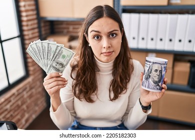 Young Hispanic Woman Working At Small Business Ecommerce Holding Money And Piggy Bank Skeptic And Nervous, Frowning Upset Because Of Problem. Negative Person. 