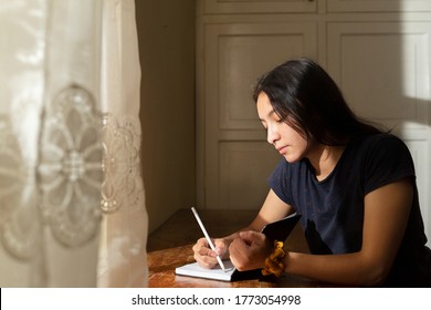 Young Hispanic Woman Writing In A Notebook, Serious Girl Writing In Her Journal In Her Room At Sunset. Concentrated Young Woman Studying At Home