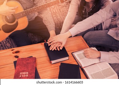 A Young Holding Guitar Joins Hand And Pray Together Over Holy Bible On Wooden Table  At Home, Christian Family, Small Group Or House Church Worship Concept