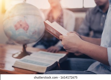 A Young  Holding And Reads Bible With His Friend Over Blurred World Globe And Open Bible On Wooden Table At Home, Christian Family, Small Group Or House Church Worship Concept, World Mission Ministry