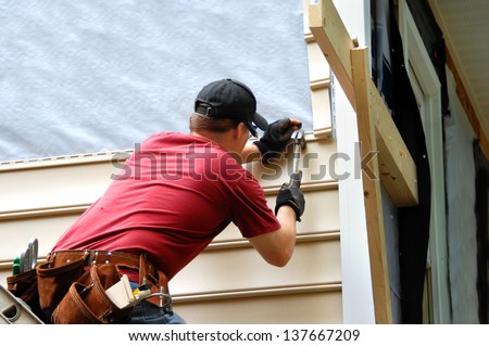 Young homeowner installs siding to his home.  He is holding a hammer and wearing a tool belt.