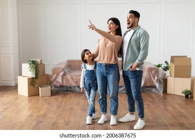 Young Homeowners Concept. Excited Young Family Of Three People Planning Interior, Decorations And Furniture In Their Own House, Standing In Empty Living Room, Woman Pointing Up At Free Copy Space