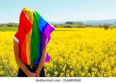 Young Homosexual Man Covered With Rainbow Gay Pride Flag, Hides His Face, In Yellow Flowers Field At Spring Sunny Day. LGBT, Homosexual Discrimination Concept.