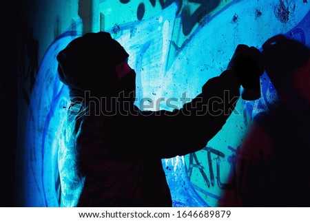 A young hooligan with a spray can stands against a concrete wall with graffiti paintings. Illegal vandalism concept. Street art