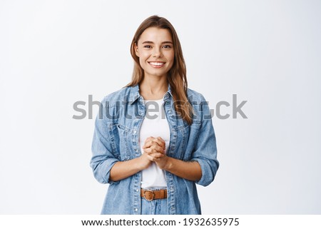 Young hopeful woman with blond hair, holding hands together and smiling politely, waiting for opportunity, ready to help client, standing against white background