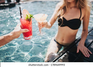 Young Hot Couple Resting At Swimpool. Cut View Of Man And Woman Cheers With Cocktails Drink In Water. Relaxing And Resting In Resort Spa. Sexy Couple With Gorgeous Body.