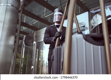 Young Hotel Employee In A Transparent Visor And Rubber Gloves Pushing A Luggage Trolley Out Of The Hotel Door.