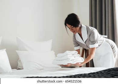 Young Hotel Maid Putting Stack Of Fresh White Bath Towels On The Bed Sheet