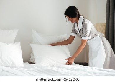 Young Hotel Maid Setting Up White Pillow On Bed Sheet In Hotel Room