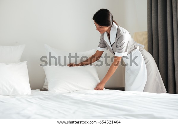 Young hotel maid setting up white pillow on bed\
sheet in hotel room