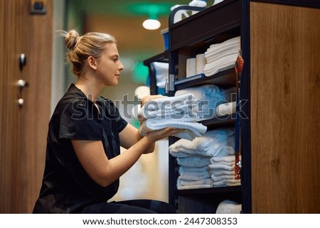Young housekeeper taking clean towels from room attendants' trolley while working in a hotel. 