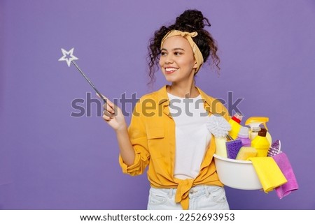 Young housekeeper woman wear yellow shirt tidy up hold basin with detergent bottles point magic wand fairy stick aside on area isolated on plain pastel light purple background studio Housework concept