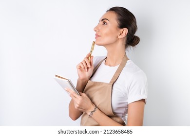 A young housewife in an apron on a white background with a notebook in her hands, chooses recipes for dinner, thinks how to please her husband, comes up with ideas for dishes