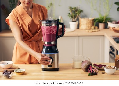 Young housewife with electric blender preparing beetroot smoothie by kitchen table with bowls containing ingredients