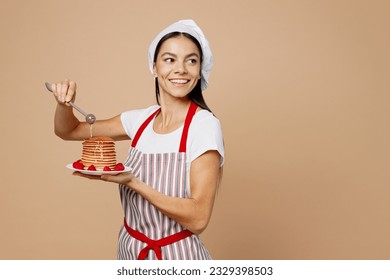 Young housewife housekeeper chef baker latin woman wear striped apron toque hat hold plate sprinkle pancakes with honey look aside isolated on plain pastel light beige background. Cook food concept - Powered by Shutterstock