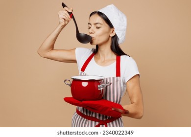 Young housewife housekeeper chef baker latin woman wear striped apron toque hat hold laddle red pot pan try dish eat with closed eyes isolated on plain pastel light beige background Cook food concept