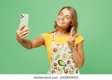 Young housewife housekeeper chef cook baker woman wear apron yellow t-shirt doing selfie shot on mobile cell phone show v-sign isolated on plain pastel green background studio. Cooking food concept