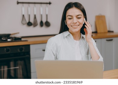 Young housewife latin IT woman wear casual clothes shirt hold use work on laptop pc computer listen music in earphones smile sit at table in light kitchen at home alone. Lifestyle cooking food concept