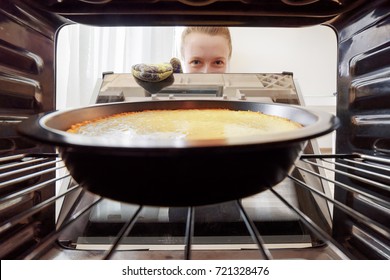 Young Housewife Looking At Cheesecake Into Oven In Kitchen. View From Inside Of The Oven. Woman Holding The Oven Door.