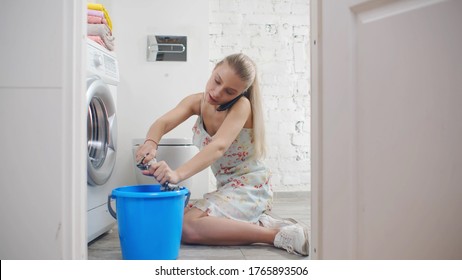 Young housewife wiping floor with cloth sitting near leaking washing machine and calling on smartphone for plumber. Woman having washer broken