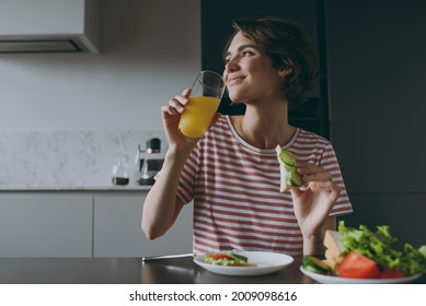 Young housewife woman 20s in casual clothes striped t-shirt eat vegeterian breakfast lunch drinking orange juice eat sandwich cooking food in light kitchen at home alone Healthy diet lifestyle concept - Shutterstock ID 2009098616