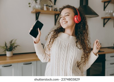 Young housewife woman of African American ethnicity in casual clothes sweater listen music in headphones use mobile cell phone stand near table in kitchen at home alone Lifestyle cooking food concept - Powered by Shutterstock