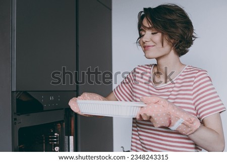 Young housewife woman in casual clothes striped t-shirt puts casserole in baking dish in oven cooking sniff delicious scent food in light kitchen at home alone Healthy diet bakery lifestyle concept.