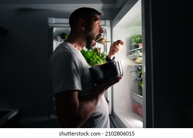 Young hungry man eating food at night and looking in open fridge. Man taking midnight snack from refrigerator - Shutterstock ID 2102806015