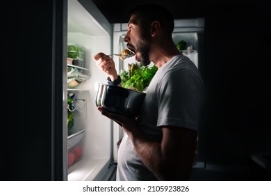 Young hungry man eating food at night from open fridge. Midnight snack and food addiction