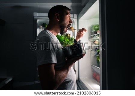 Young hungry man eating food at night and looking in open fridge. Man taking midnight snack from refrigerator