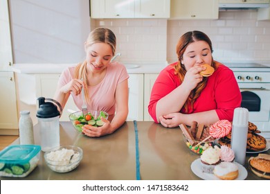 Young Hungry Slim And Overweight Women In Kitchen Eating Food. Healthy And Unhealthy Meal. Salad Vs Burger. Body Positive. Healthy Lifestyle.