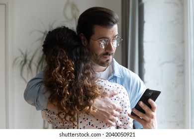 Young husband hugs wife holds cellphone check messages in social media suspect her in treason. Broken trust in relationships, jealous boyfriend, mistrust in relations, crisis of family life concept