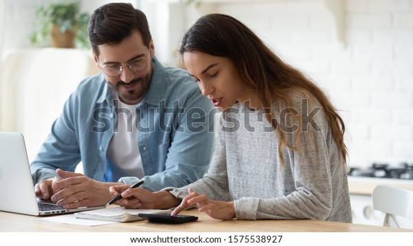 Young husband and wife using calculator laptop\
computer manage finances calculate bills tax talk doing paperwork\
together sit at home table discuss family mortgage loan money\
payment planning budget