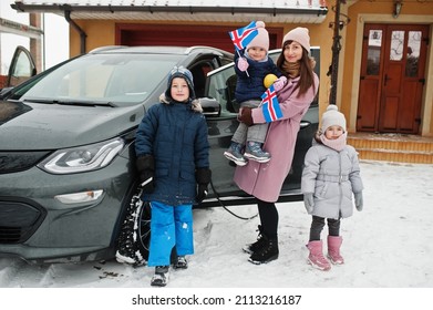 Young Icelandic Mother With Kids Hold Iceland Flags And Charging Electric Car In The Yard Of Her House At Winter.