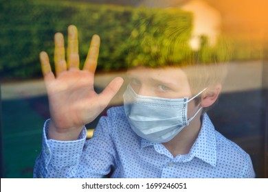 Young Ill Boy Looking Sad With Protective Mask At Home Behind Window In Quarantine And Lockdown Missing School And Freedom During Covid-19 Coronavirus Worldwide Pandemic 